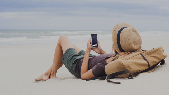 Yngre kvinne som ligger på stranden med sin mobil.